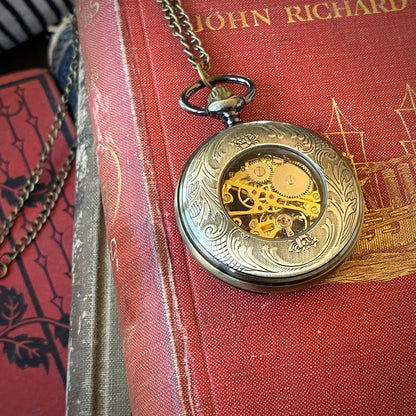 Antiqued brass Zodiac mechanical pocket watch face down to display an ornate back panel with a window to internal gears in the center.