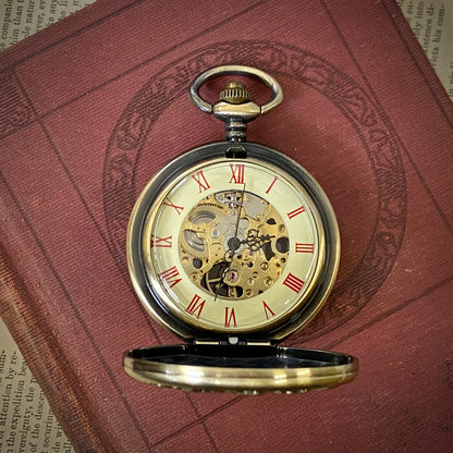 Antiqued brass Zodiac mechanical pocket watch. The watch is displayed open, with red roman numerals on a cream-colored boarder. Watch internal gears are visible in the center of the watch.