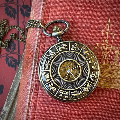 Antiqued brass mechanical pocket watch with zodiac symbols around the edge of the lid. The watch is closed, but some of the internal gears are visible through the lid. The watch is on a necklace chain.