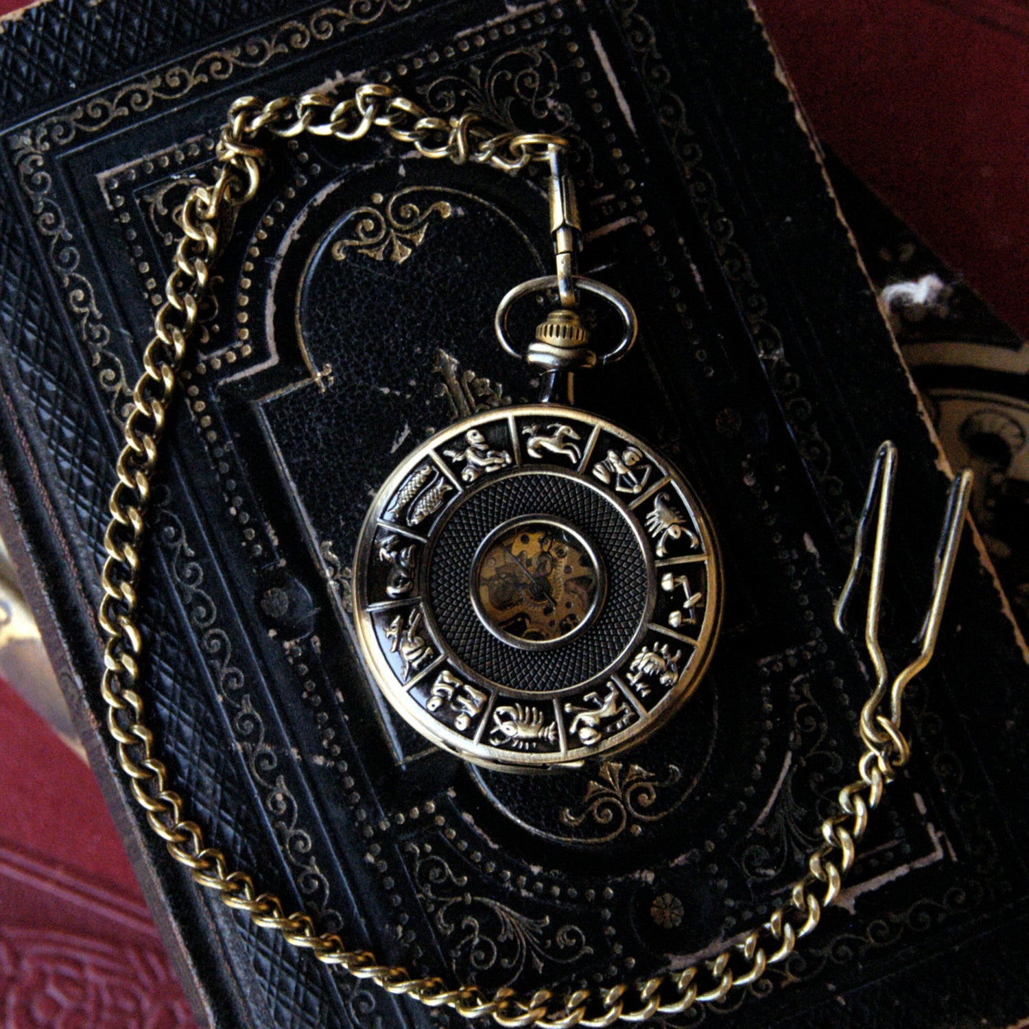 Antiqued brass mechanical pocket watch with zodiac symbols around the edge of the lid. The watch is closed, but some of the internal gears are visible through the lid. This watch is on a pocket chain.