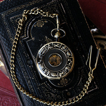 Antiqued brass mechanical pocket watch with zodiac symbols around the edge of the lid. The watch is closed, but some of the internal gears are visible through the lid. This watch is on a pocket chain.