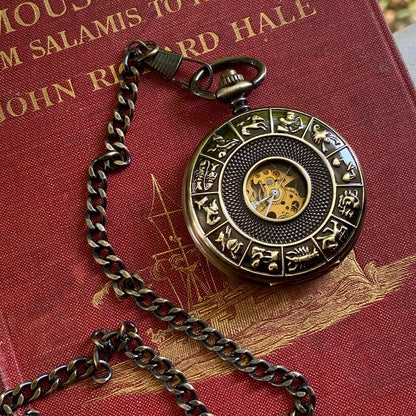 Antiqued brass mechanical pocket watch with zodiac symbols around the lid. The watch is closed, but some of the internal gears are visible through the lid.