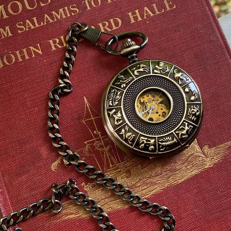 Antiqued brass mechanical pocket watch with zodiac symbols around the lid. The watch is closed, but some of the internal gears are visible through the lid.