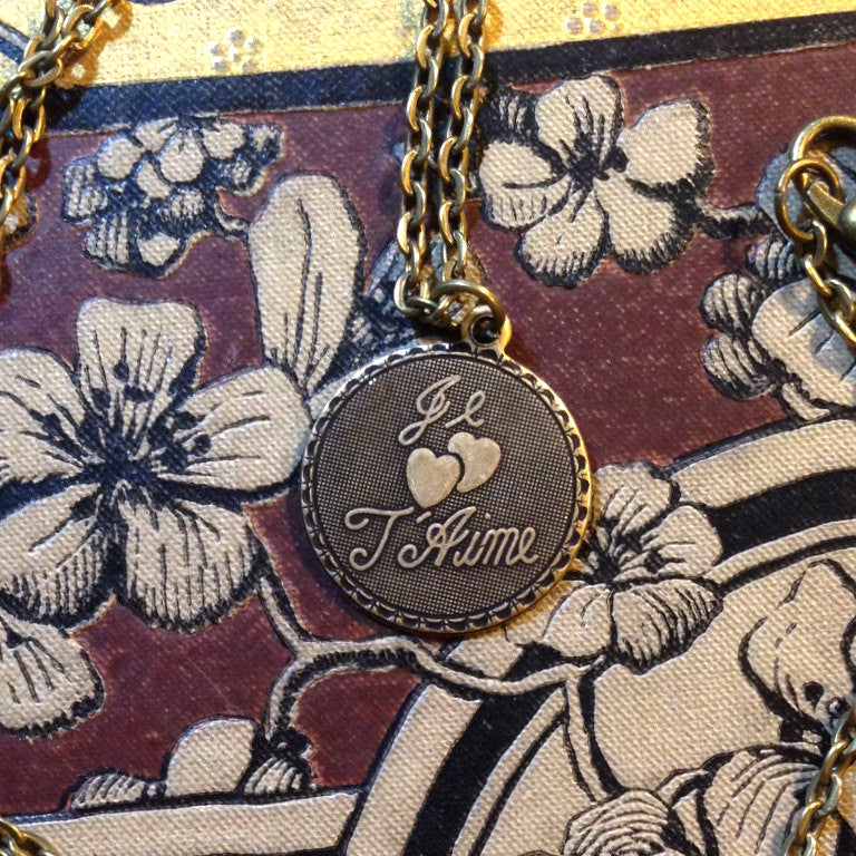 The phrase 'Je T'Aime' and two hearts on a circular brass pendant on a brass chain.