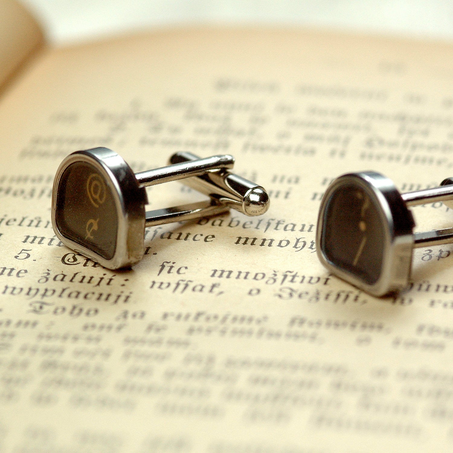 A pair of silver cufflinks with typewriter keys attached to them. Typewriter keys are metal-rimmed and tombstone-shaped with white letters on a black background.