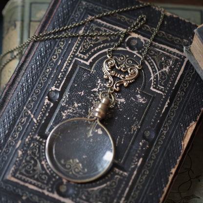 Antiqued brass round-lens glass magnifying glass attached to an angel wings connector and a necklace chain.