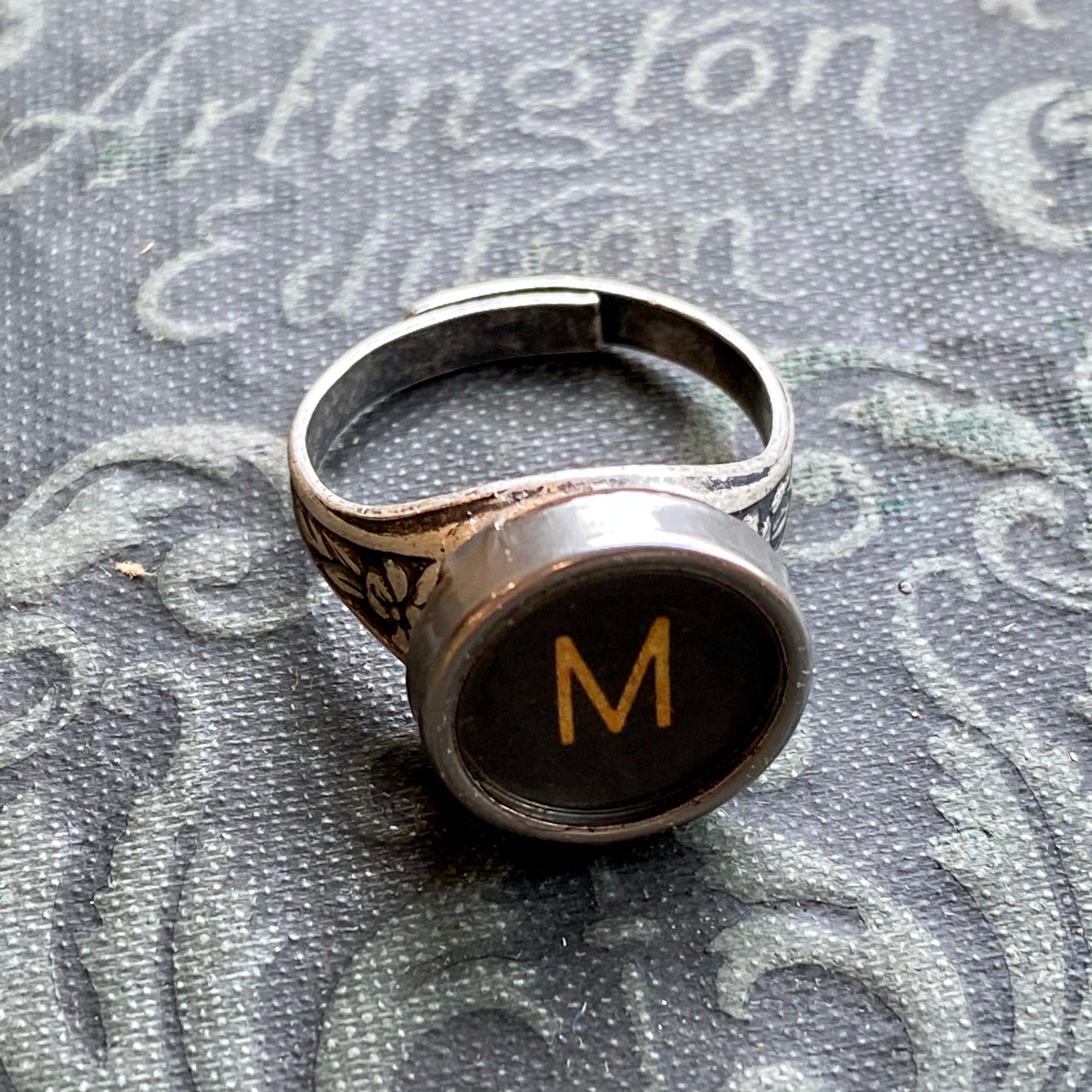 Silver adjustable ring with an authentic vintage typewriter key as the centerpiece. The key is metal-rimmed with glass covering an off-white letter 'M' on a black background.